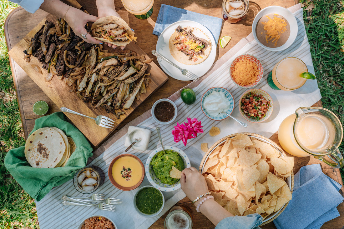 Food display with guacamole, chips, someone making a fajita, spicy sliced beef, queso, and a margarita pitcher.
