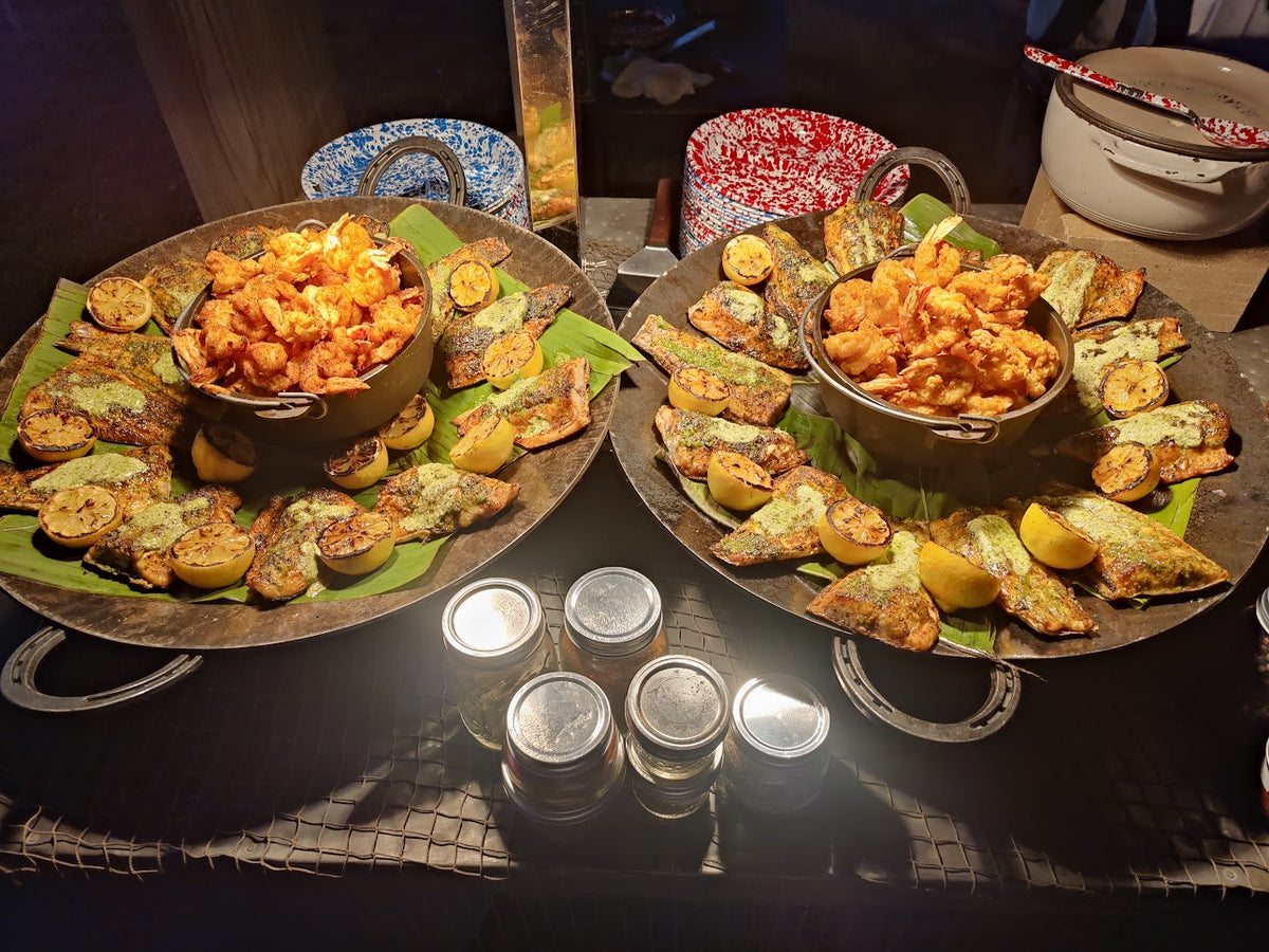 grilled catfish in two display dishes under a heat lamp with sliced lemons and fried onions in a bowl.