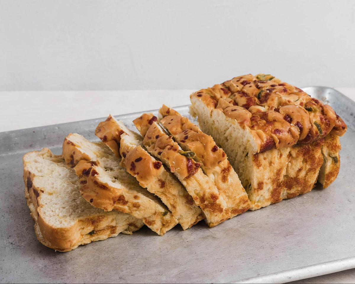 Sliced artisanal homemade bread with a golden-brown baked crust and visible jalapeño pieces, arranged on a metal baking sheet against a light neutral background.