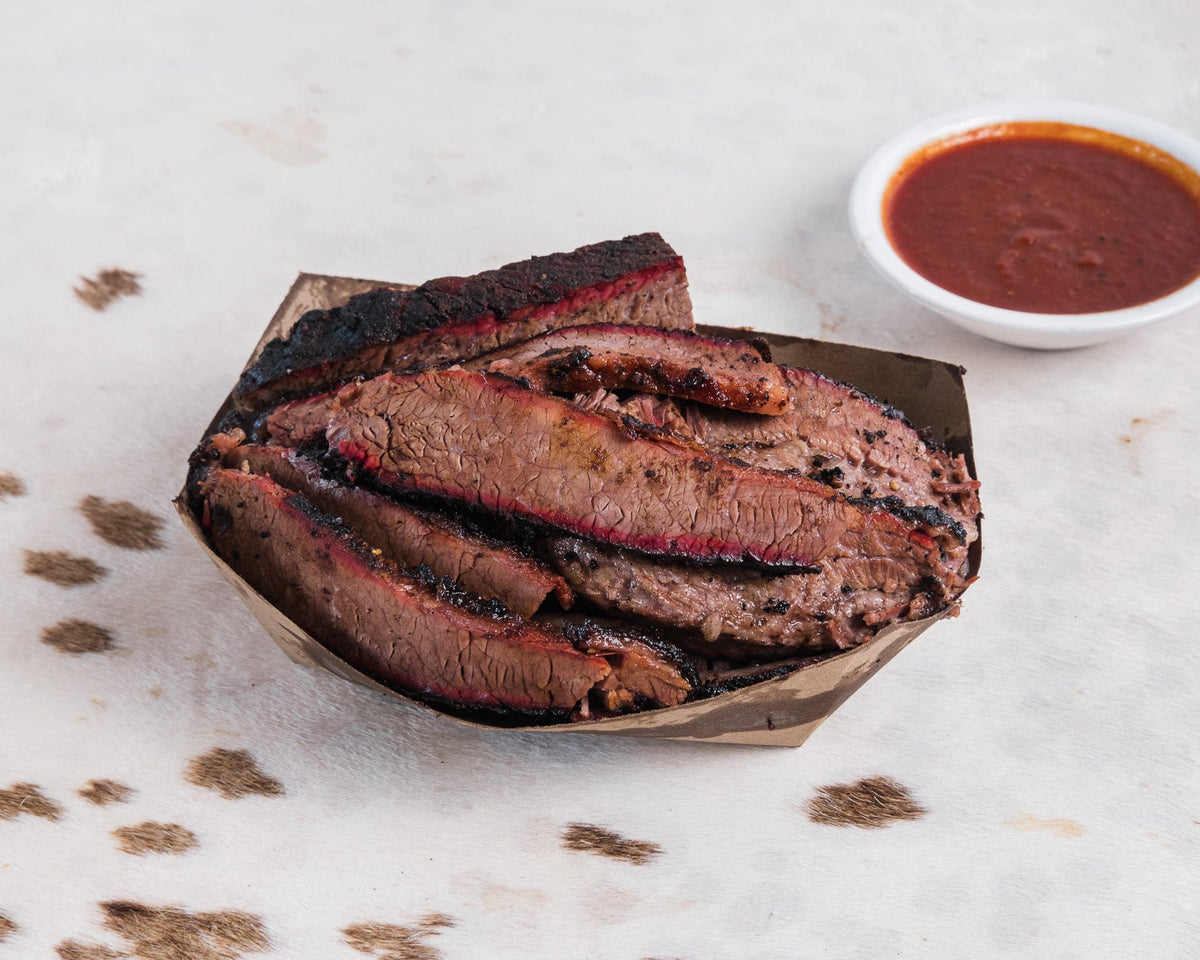 Slices of smoked brisket with charred edges served on a paper tray, accompanied by a small white bowl of red barbecue sauce on a light stone surface.