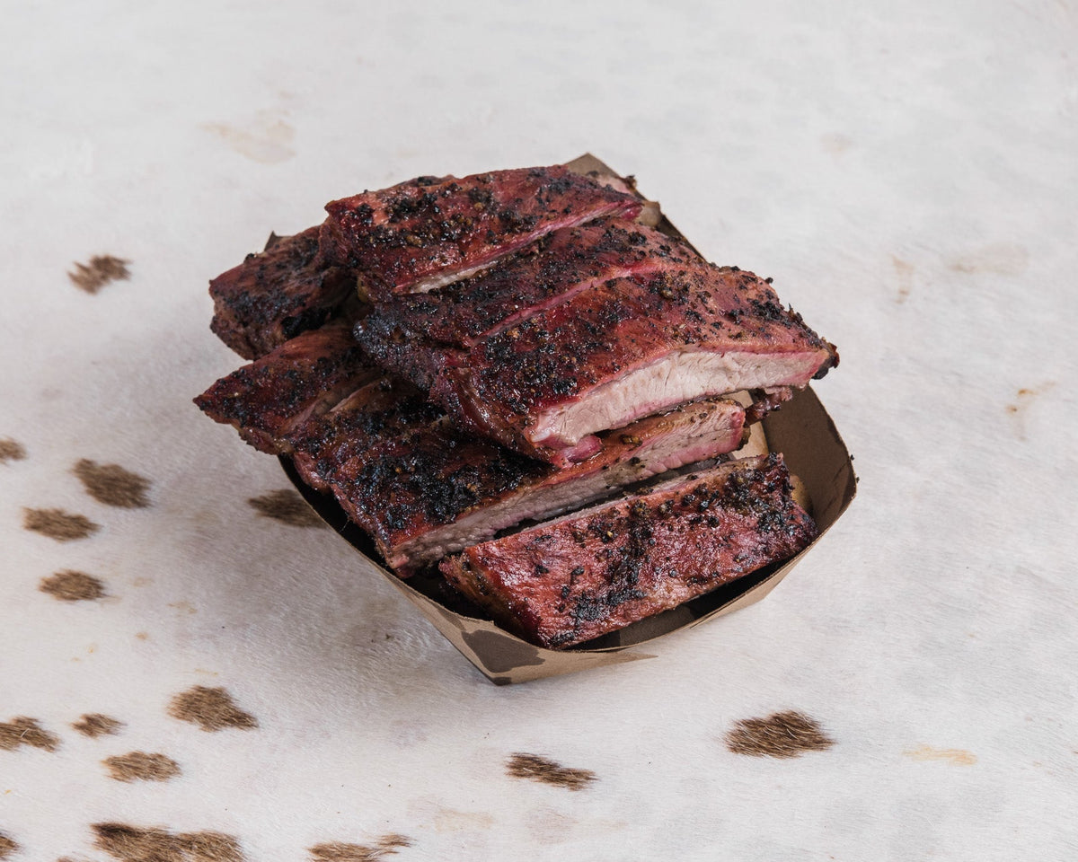 Grilled barbecue ribs stacked in a small paper tray on a light textured surface with scattered seasoning specks.
