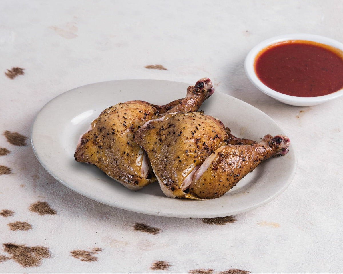 Two halves of a cooked chicken leg with golden-brown skin on a white oval plate, served with a small white bowl of red dipping sauce on a light textured surface.