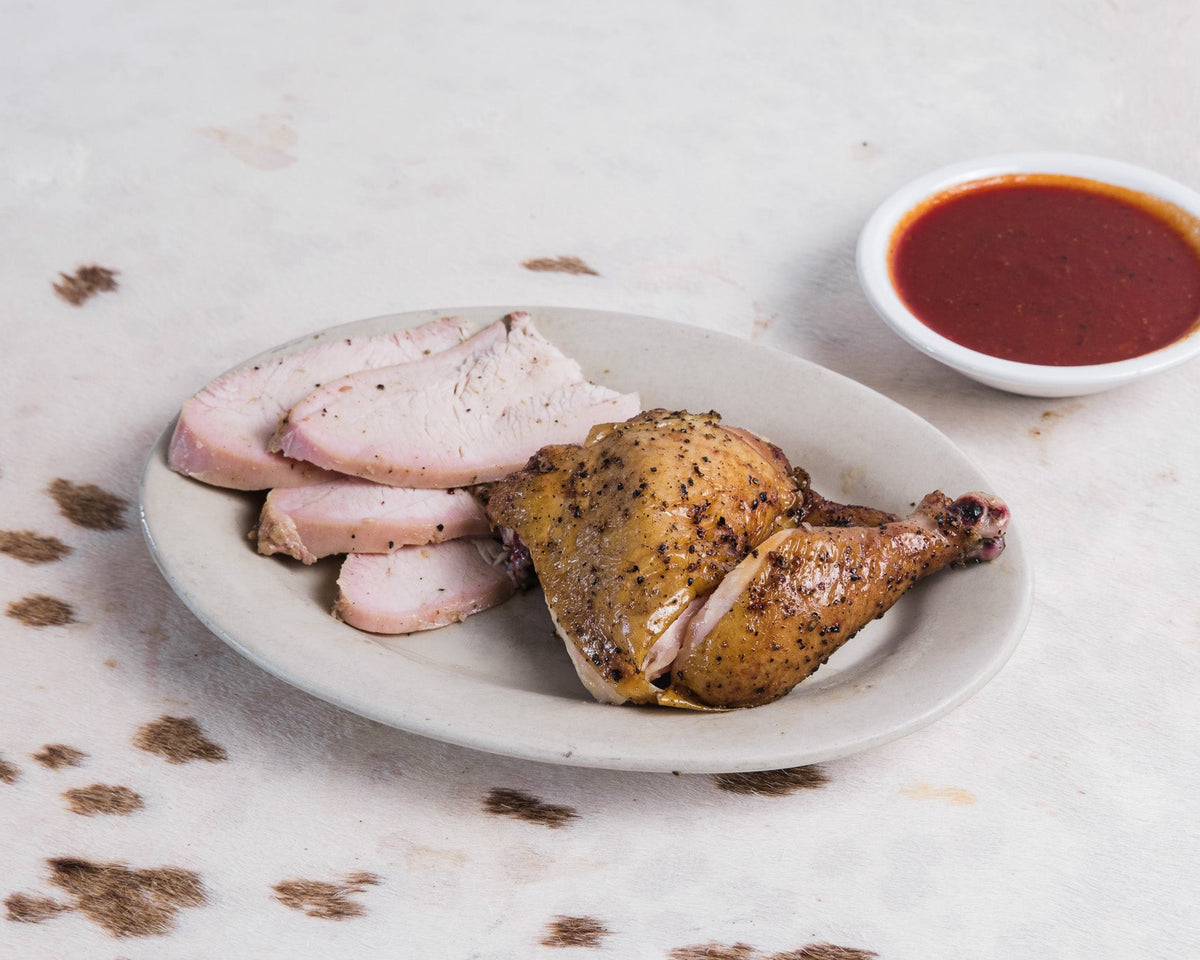 A plated meal featuring a roasted chicken leg with a small white bowl of red bbq sauce on a light textured surface.