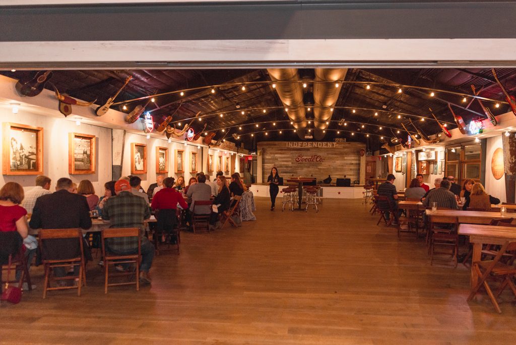 people eating around tables in a Goode Co. dining hall, and a woman returning to her table with a full plate.