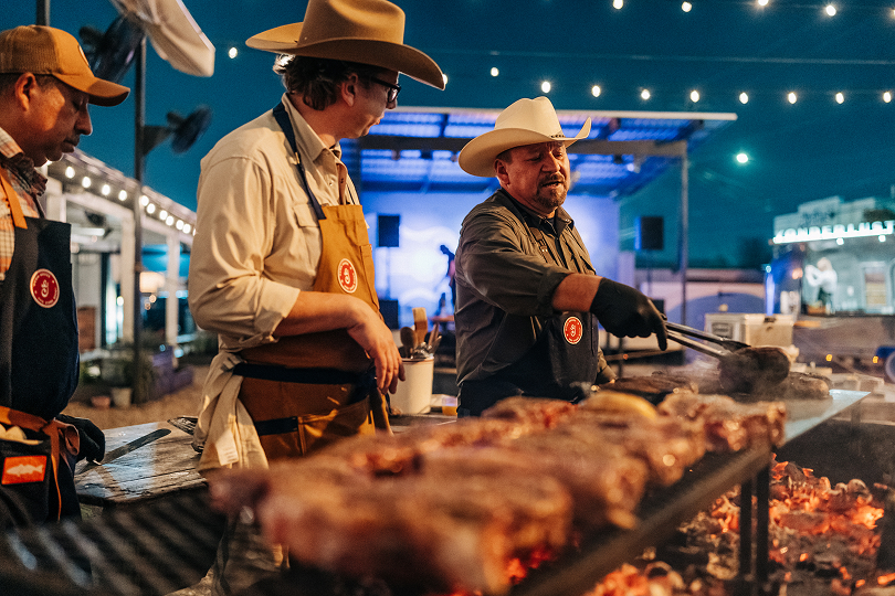 Three Stetson-wearing cowboys barbequing steaks on a large grill. 
