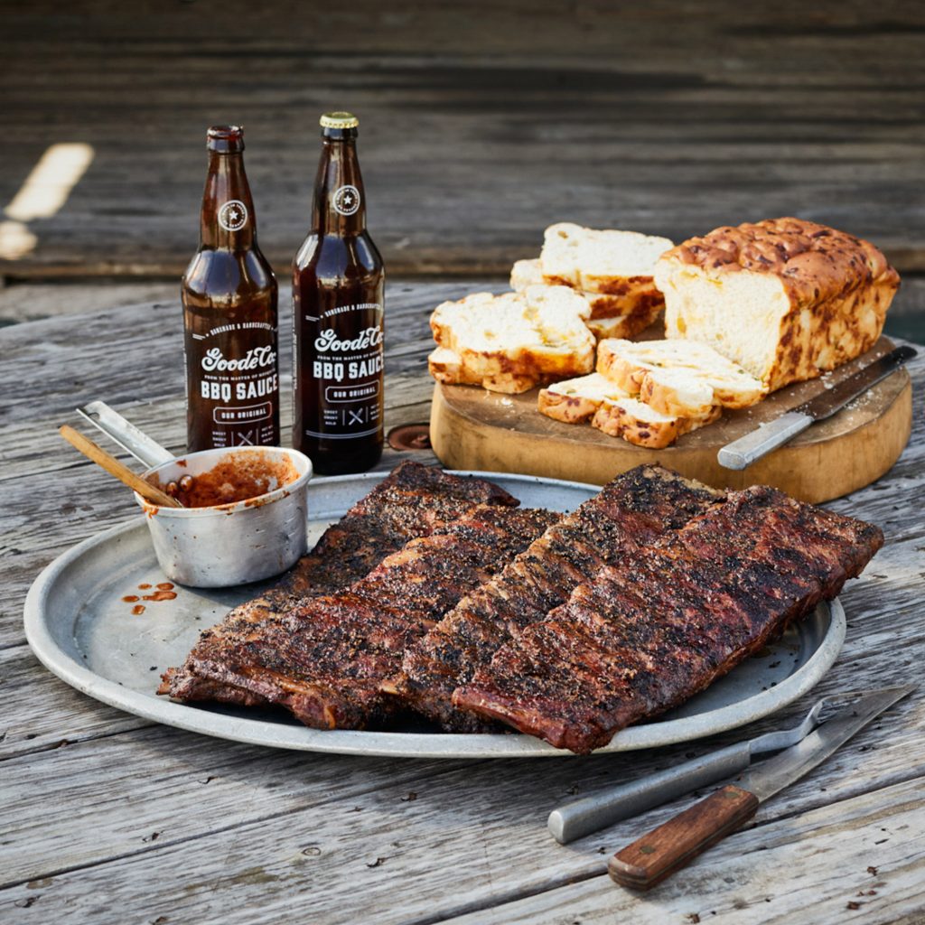 Fresh BBQ’d ribs on a platter with sauce, fork and knife, and sliced cheese bread in the background.