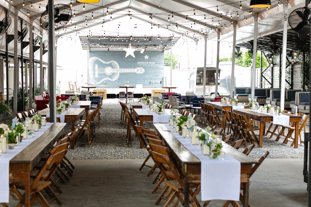 Panoramic shot of long wooden tables with white linen table runners set with candles and flowers in vases and a stage at the back of the room.