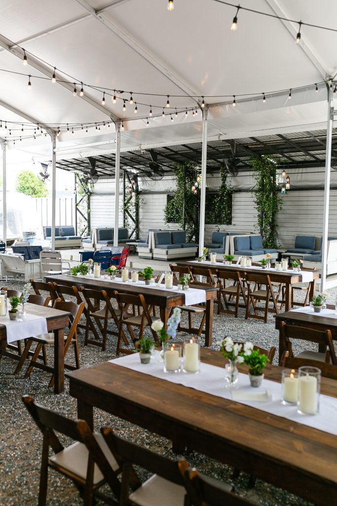 Long wooden tables with white table runners, lit candles and bouquets of flowers, and blue fabric booths in the corner with white patio lights strung up on the ceiling.