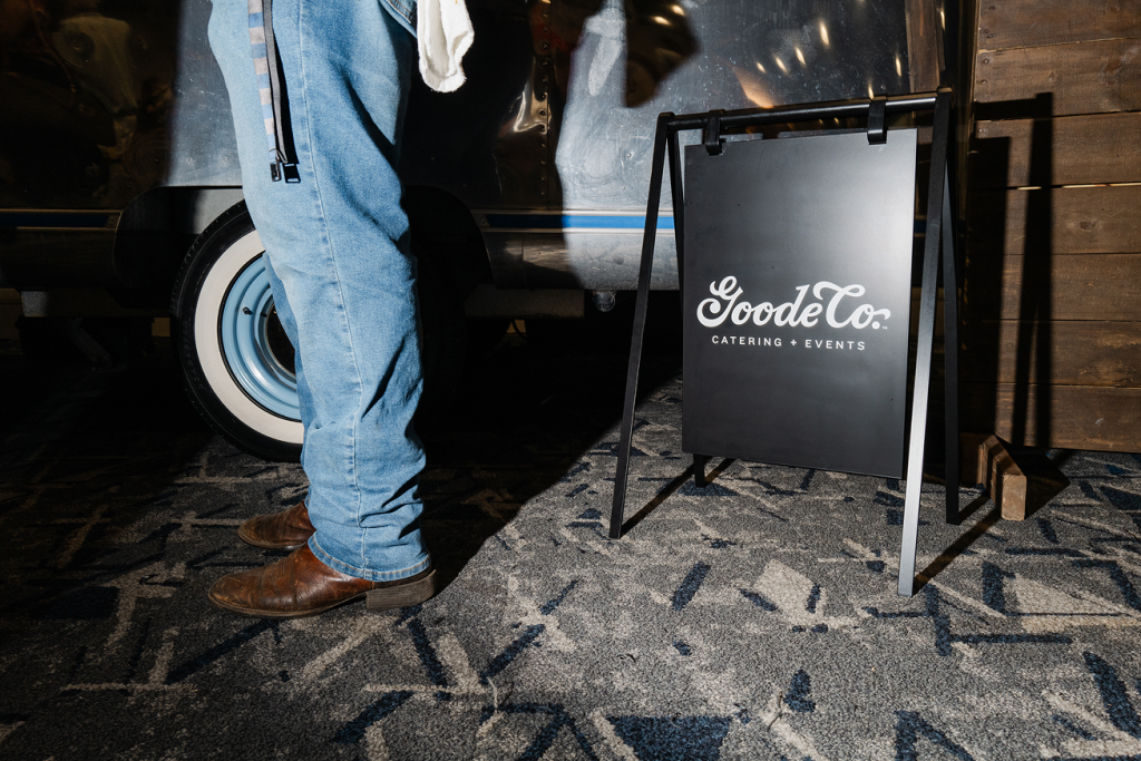 Goode Co. events & catering sign in black with a jean-clad cowboy boot-wearing man standing next to it.

