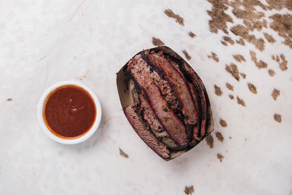 Sliced brisket in a tiny cardboard serving dish with a side of sauce.