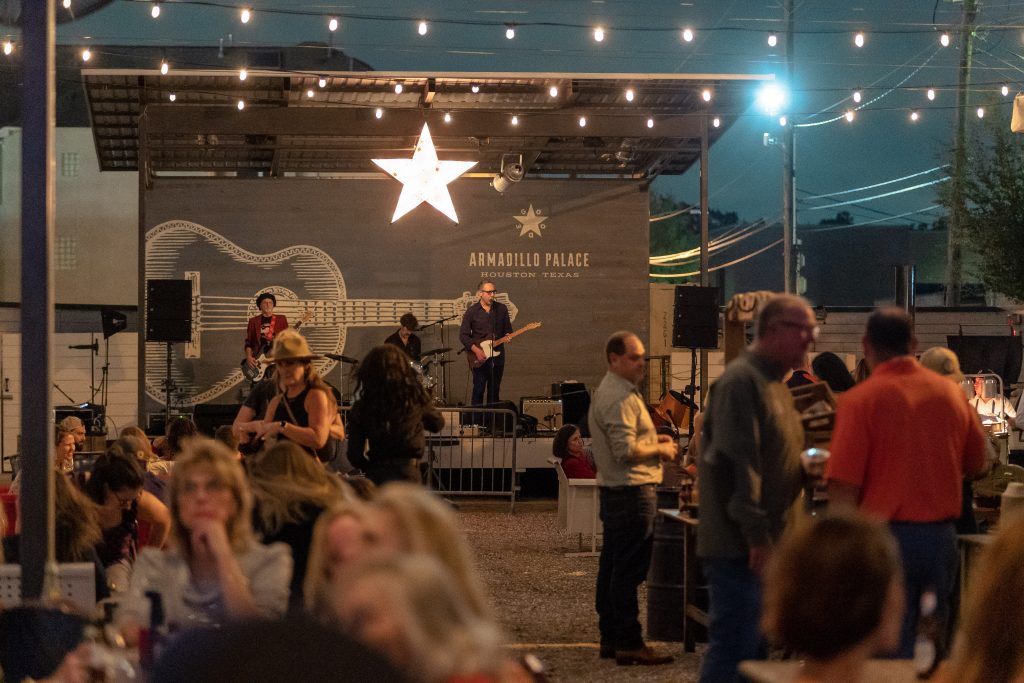 Wide shot of the Armadillo Palace event stage outside, with someone playing on guitar and others standing around and listening to the music