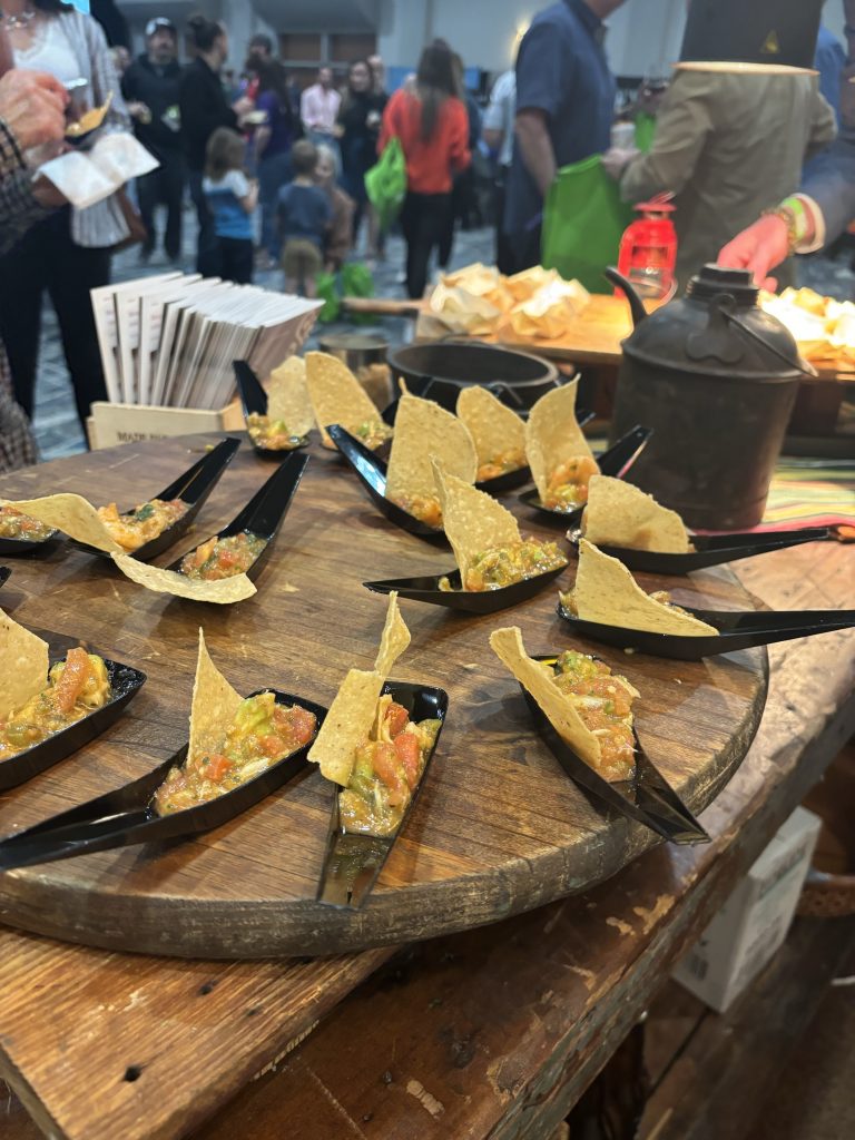 Wooden cutting board featuring serving boat spoons, each with a tortilla chip and salsa.