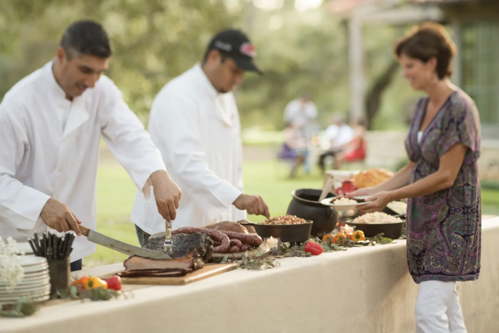 Two men dressed in culinary white smocks, carving beef and serving food on a buffet line while a customer waits.