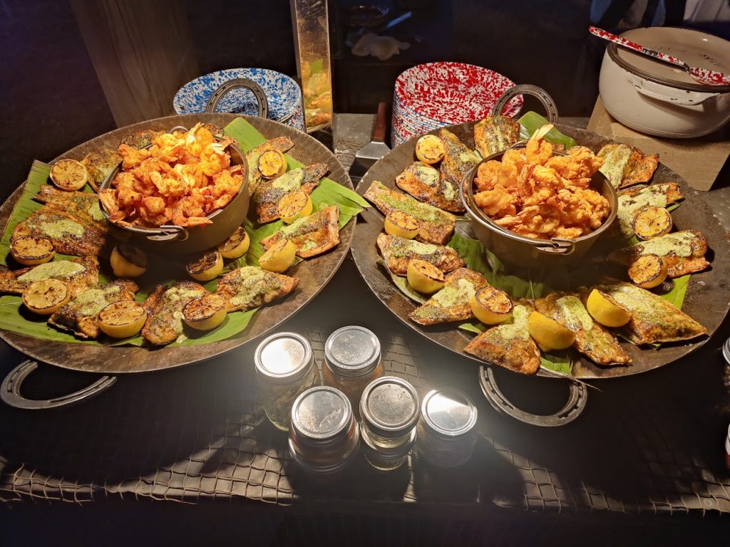 grilled catfish in two display dishes under a heat lamp with sliced lemons and fried onions in a bowl.